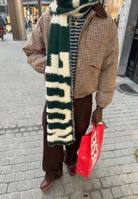 Chaqueta marrón de estampado pata de gallo con cuello marrón, bufanda con patrón verde y blanco, camisa a rayas y pantalones marrón oscuro, sosteniendo un bolso rojo de Diesel.