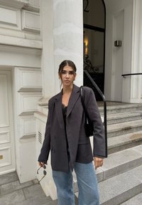 Szary oversize blazer o podwójnym zapięciu, w połączeniu z czarną koronkową bluzką i szerokimi, niebieskimi dżinsami, trzymający okulary przeciwsłoneczne.