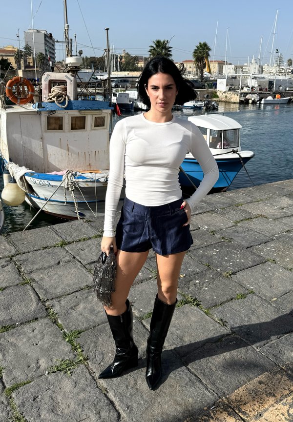 Vrouw met zwart bobkapsel, witte top met lange mouwen, marineblauwe shorts en zwarte laarzen staand op een stenen pier bij aangemeerde boten in fel daglicht.