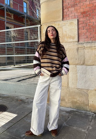 Suéter de punto a rayas en marrón, crema y rosa; pantalones anchos blancos; zapatos de plataforma marrones; en un entorno urbano al aire libre.