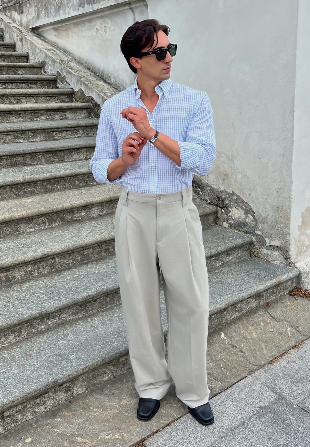 Camicia a quadri azzurro chiaro, pantaloni beige a gamba larga, scarpe nere e occhiali da sole; in piedi su gradini di pietra contro un muro.