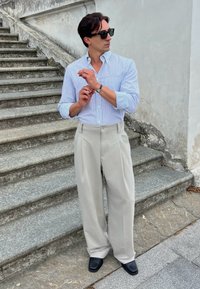 Camicia a quadri azzurro chiaro, pantaloni beige a gamba larga, scarpe nere e occhiali da sole; in piedi su gradini di pietra contro un muro.
