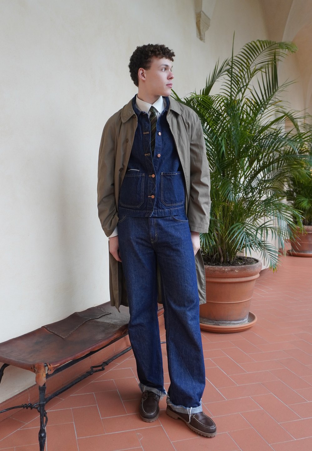 Καραμέλα παλτό πάνω από ένα σκούρο τζιν μπουφάν και παντελόνι, συνδυασμένο με καφέ δερμάτινα παπούτ�σια. Εσωτερικός χώρος με γλάστρες και πλακόστρωτο δάπεδο.