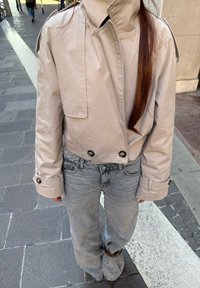 Světlá šedá zkrácená bunda s velkými knoflíky, dvěma předními kapsami a vysokým límcem, doplněná o vybledlé šedé džíny. Hladká textilní textura.