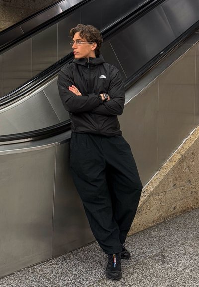 Chaqueta negra con cremallera y logo, pantalones sueltos negros y zapatos negros. De pie cerca de una escalera mecánica en un entorno moderno.