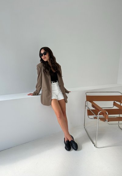 Blazer marrón sobre una blusa negra, pantalones cortos blancos y mocasines negros. El entorno presenta una pared blanca y una silla de cuero marrón.