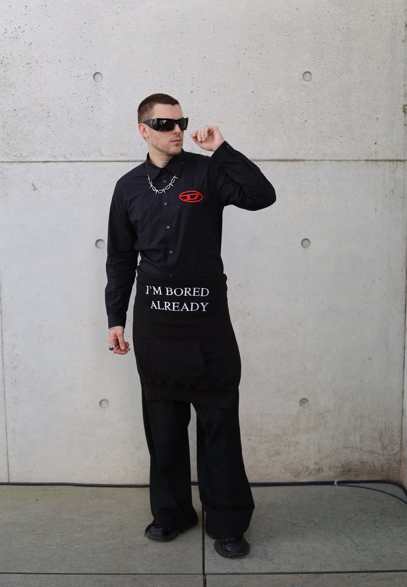 Μαύρο πουκάμισο με μακριά μανίκια και κόκκινο λογότυπο, μαύρη ποδιά με λευκό κείμενο "ΕΙΜΑΙ ΗΔΗ ΒΑΡΙΕΜΑΙ", φαρδύ μαύρο παντελόνι και σκούρα γυαλιά ηλίου με φαρδύ κολιέ.
