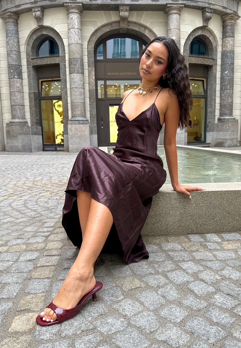 Vestito in satin di un profondo viola con spalline sottili e uno spacco alto; sandali con tacco a scivolo in pelle verniciata rossa; texture liscia e lucida.
