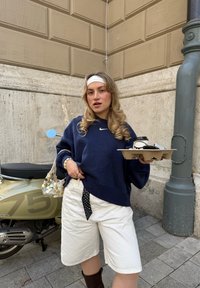 Felpa Nike blu navy, pantaloni corti bianchi, cintura a pois, che tiene un vassoio per tazze di caffè; sfondo con un monopattino chiaro e un muro di pietra.