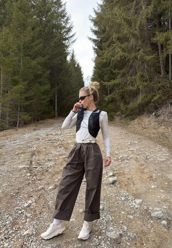 Žena u sunčanim naočalama pije iz prsluka za hidrataciju dok stoji na kamenitoj šumskoj stazi okružena visokim zimzelenim drvećem.