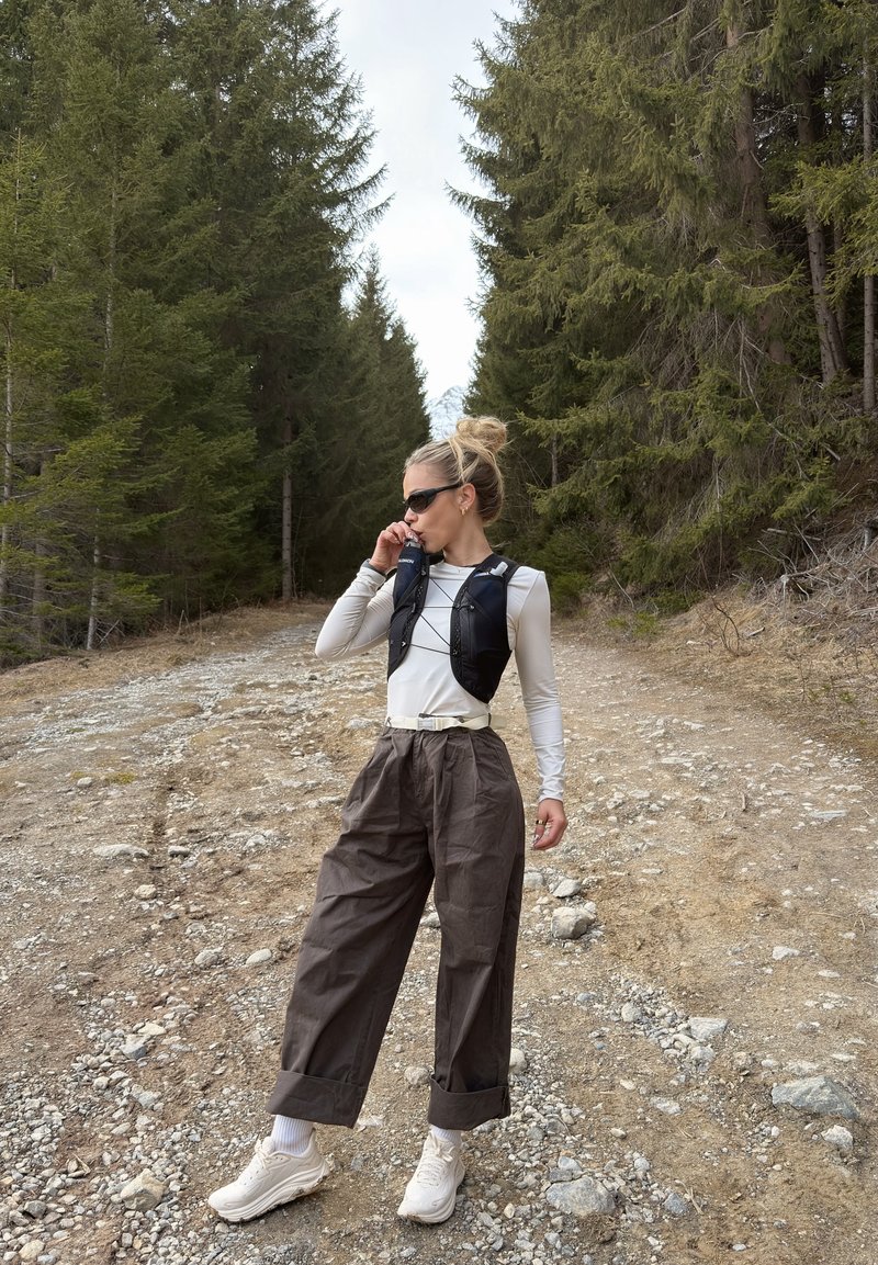 Ženska z sončnimi očali pije iz hidracijskega jopiča, medtem ko stoji na skalnati gozdni poti obkroženi z visokimi zimzelenimi drevesi.