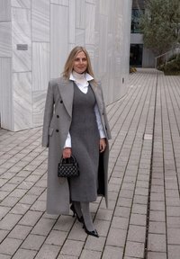 Sivý vlnený kabát s čiernymi gombíkmi, vrstvený cez sivé šaty a bielu košeľu. Čierna prešívaná kabelka a špicaté podpätky dopĺňajú vzhľad.