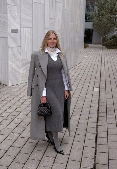 Abrigo gris de lana con botones negros, superpuesto sobre un vestido gris y una camisa blanca. Bolso negro acolchado y tacones puntiagudos completan el conjunto.