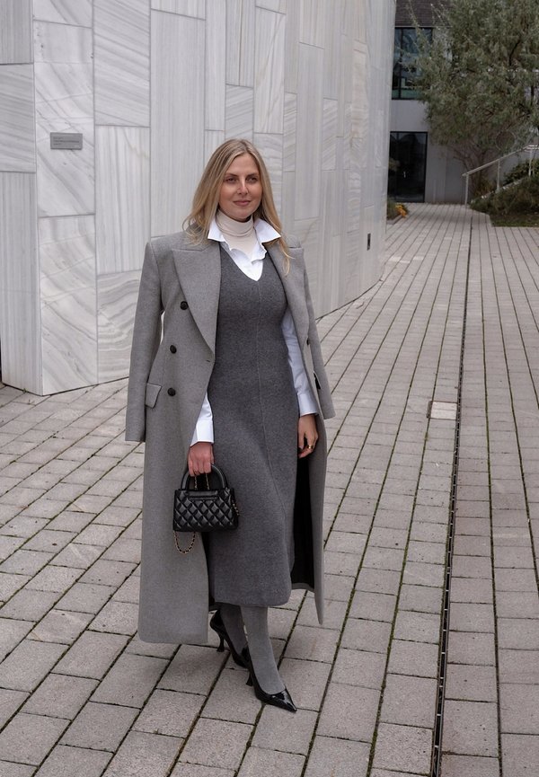 Grå ullfrakk med svarte knapper, lagvis over en grå kjole og hvit skjorte. Svart quiltet håndveske og spisse hæler fullfører antrekket.