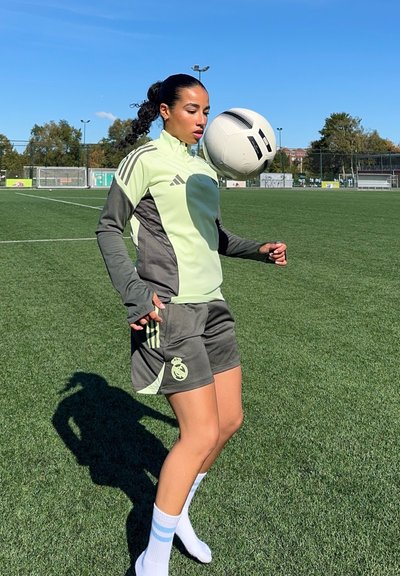 Ropa deportiva verde y gris con tres rayas; balón de fútbol equilibrado en la frente; fondo de campo de hierba; día soleado.