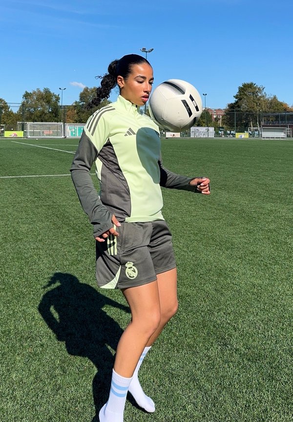 Tenue de sport verte et grise avec trois rayures ; balle de football équilibrée sur le front ; arrière-plan de terrain de gazon ; jour ensoleillé.