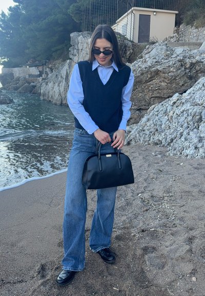 Mujer con gafas de sol, camisa azul, chaleco negro y vaqueros sostiene un bolso negro parada en una playa rocosa cerca del agua.