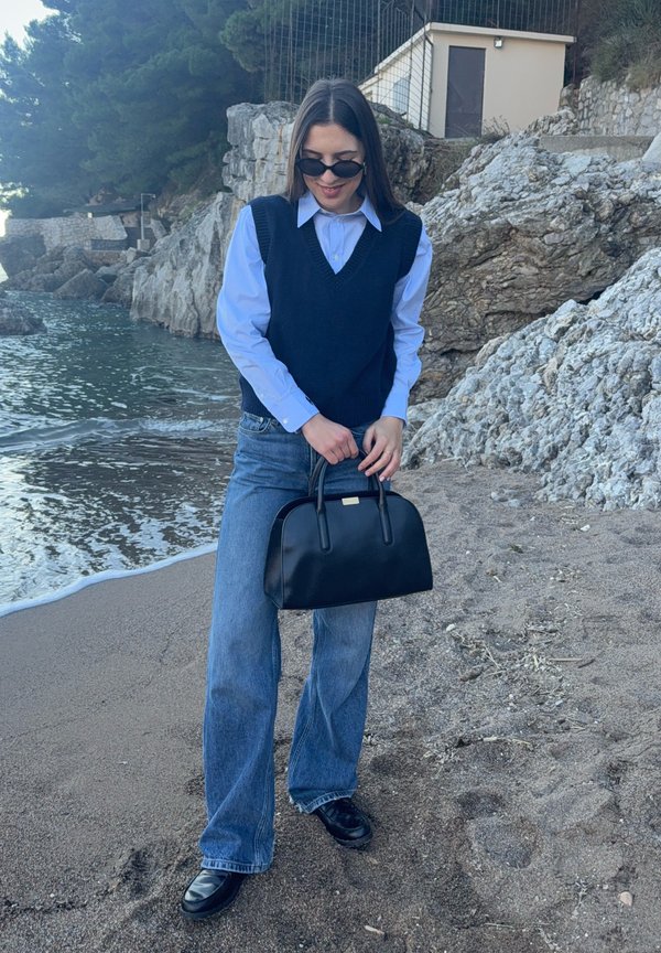 Mujer con gafas de sol, camisa azul, chaleco negro y vaqueros sostiene un bolso negro parada en una playa rocosa cerca del agua.