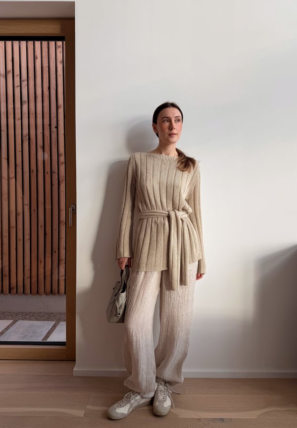 Vrouw staande binnenshuis, gekleed in een beige geribbelde trui met riem, losse crèmekleurige broek, witte sneakers, en houdend een kleine lichtgrijze handtas vast.