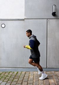 Vyras pilkai geltonoje striukėje bėgioja akmenine grindinio taku šalia pilkos betono sienos su dviem sieniniais šviestuvais.