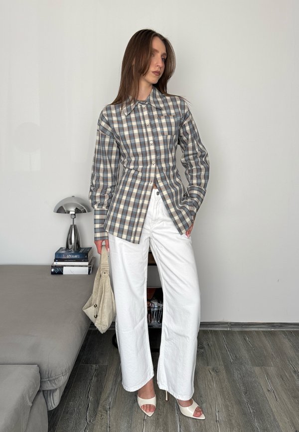 Femme portant une chemise à manches longues à carreaux, pantalon blanc à jambes larges, talons blancs ouverts à l’avant, tenant un sac à main beige, debout à l’intérieur près d’un canapé.