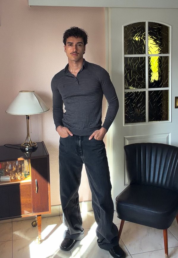 Homme aux cheveux bouclés et à la moustache portant une chemise sombre à manches longues et un jean noir se tient à l'intérieur près d'une lampe, d'une armoire et d'une chaise noire.