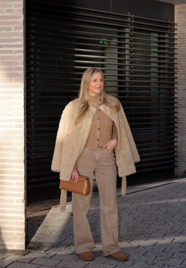 Donna che indossa un cappotto di pelliccia beige appoggiato sulle spalle, maglione beige lavorato a trecce, pantaloni a gamba larga, che tiene una pochette marrone, in piedi su un marciapiede lastricato.