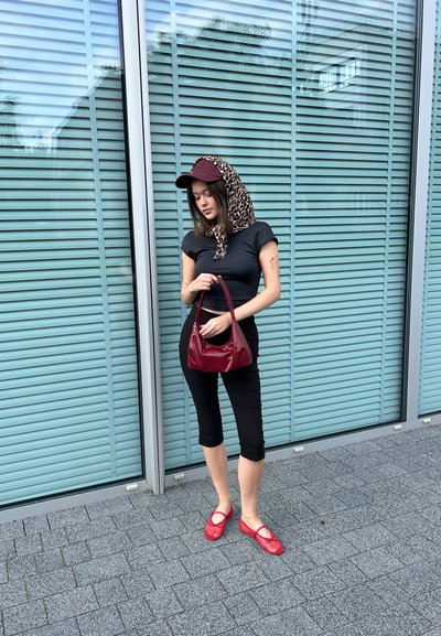 Top corto negro, pantalones capri negros, zapatos de rejilla rojos, bolso burdeos. Pañuelo en la cabeza con estampado de leopardo, gorra burdeos, de pie junto a un edificio de cristal.