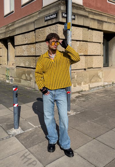 Suéter a rayas amarillas y negras, vaqueros azules holgados, botas de plataforma negras y guantes sin dedos. Los accesorios incluyen gafas de sol y joyería.