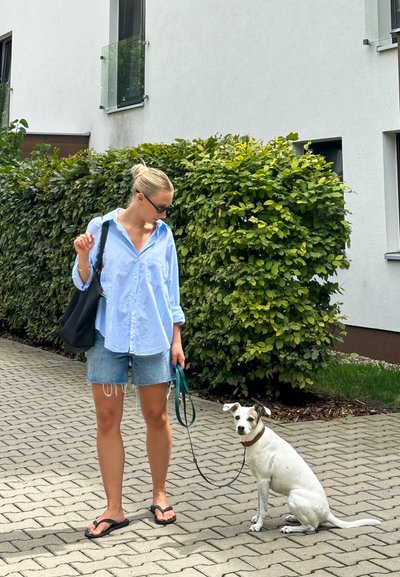 Camisa de botón azul claro, pantalones cortos de mezclilla, chanclas negras. Bolsa de mano negra. Perro blanco con detalles marrones, sentado, con una correa verde. Fondo de seto verde.