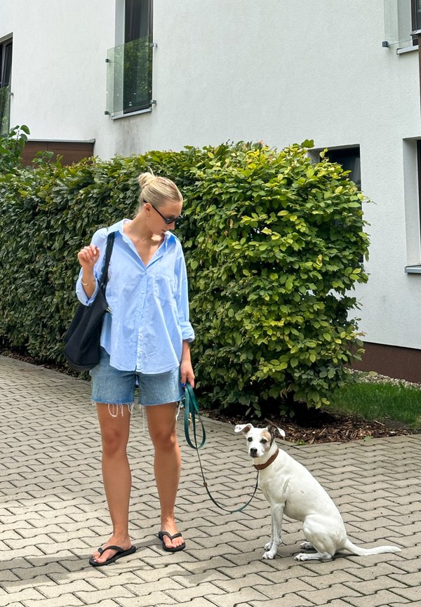 Svetlo modra srajca s kratkimi rokavi, jeans kratke hlače, črne japonke. Črna torba za na ramo. Bela pes s rjavimi akcenti, sedi, nosi zeleno povodec. Zelen živec v ozadju.