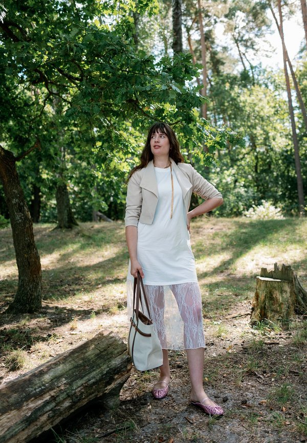 Femme en robe blanche et veste beige, debout sous un arbre dans la forêt, tenant un sac à main beige et regardant sur le côté.