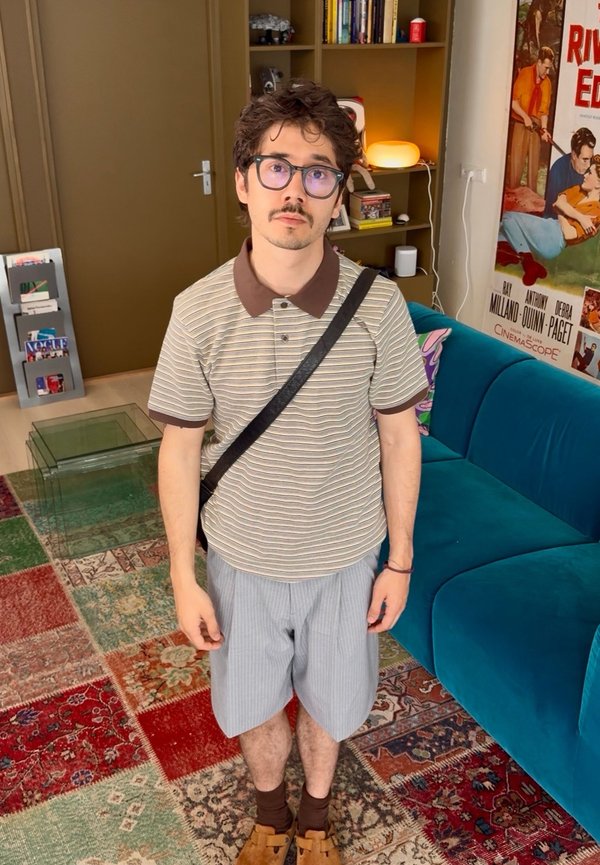Ung mand med briller iført en stribet polo, shorts og skuldertaske står i et farverigt stue med bogreol, blå sofa og vintageplakat.