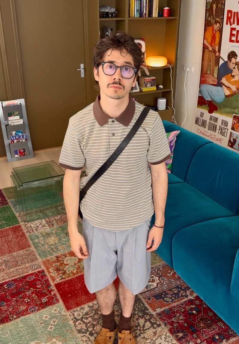 Jeune homme avec des lunettes portant un polo rayé, un short et un sac à bandoulière, debout dans un salon coloré avec une bibliothèque, un canapé bleu et un poster vintage.