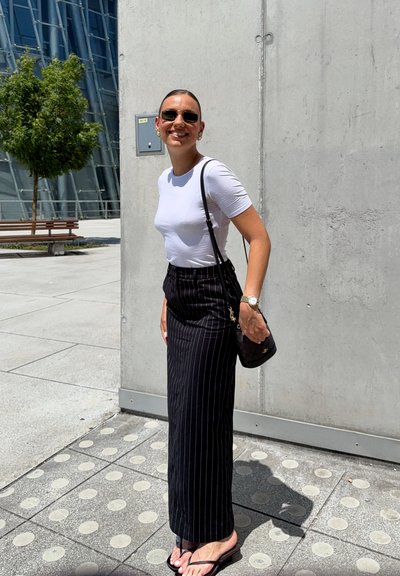 Falda a rayas negras, camisa blanca ajustada, bolsa negra y chanclas, con un fondo de una pared de concreto y arquitectura moderna.