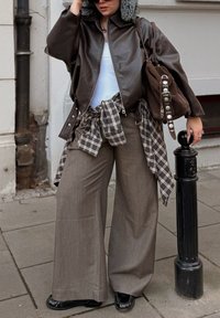 Giacca di pelle marrone con cappuccio foderato in pelliccia, canottiera bianca, pantaloni larghi a quadri grigi, borsa marrone con dettagli argento e scarpe nere.