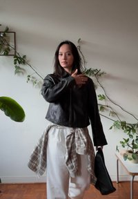 Giacca nera in pelle crop con colletto alto, indossata sopra una camicia bianca annodata con una flanella a quadretti. Il modello tiene una borsa nera. Foliaggio verde sullo sfondo.