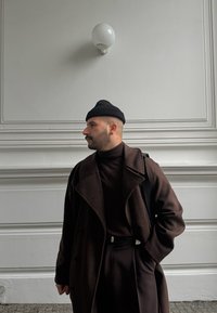 Καφέ μάλλινο παλτό πάνω από ένα σκούρο ζιβάγκο, συνοδευόμενο από μία μαύρη σκουφίτσα. Σε φόντο μία λευκή τοιχοποιία με τοίχου φωτιστικό.