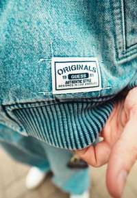 Jeans jakna v svetlo modri barvi z rebrastimi manšetami; oznaka pravi "ORIGINALS 1981 GUESS AVTENTIČEN STIL KONCIPIRAN V LOS ANGELESU."