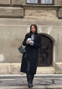 Črn volnen plašč, sivi pleteni šal, puhaste sive rokavice, črna torbica s kovinsko verižico, črni škornji do kolen, v roki drži skodelico kave, na ozadju kamniti zid.