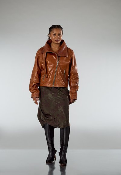 Chaqueta de cuero marrón con cuello sobredimensionado, cierre de cremallera y detalles sutiles. Combinada con una falda verde oscuro y botas negras hasta la rodilla.