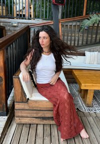 Bijela majica bez rukava, duga crvena teksturirana suknja, drveno sjedište s tkanim pokrivačem, opušteno vanjsko okruženje s zelenilom i drvenim ukrasima.