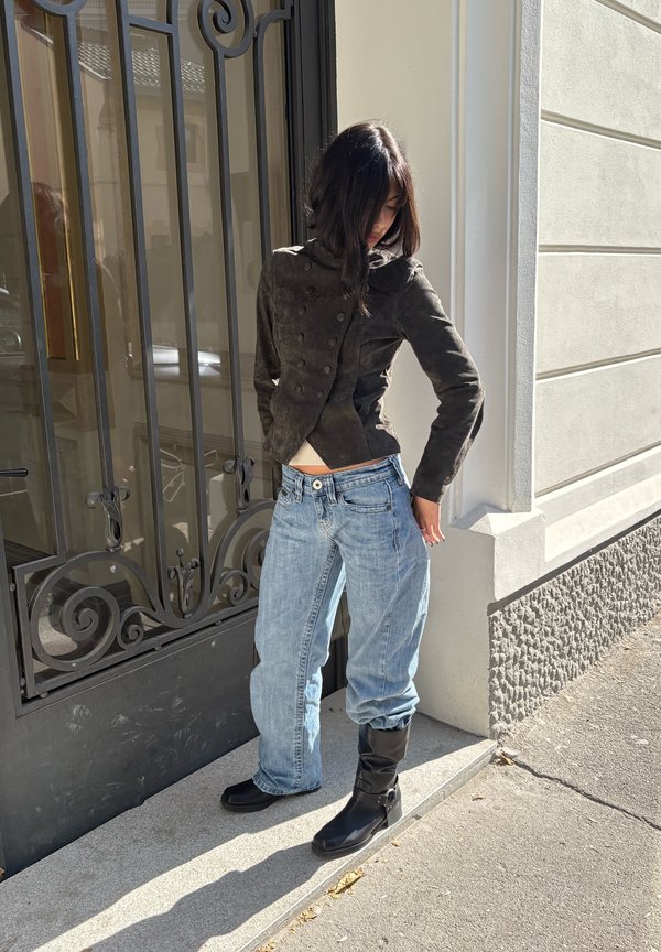 Una mujer posa al sol junto a una verja de hierro ornamentada. Lleva una chaqueta oscura, unos vaqueros azules holgados y botas negras.
