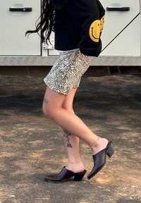 Sötétbarna tűsarkú mules sima felülettel, ívelt formával és nyitott hátsó résszel, mintás rövidnadrággal párosítva, amelyen láb tetoválások láthatók.