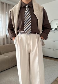 Osoba w brązowej koszuli, w paski krawacie, beżowych spodniach i swetrze zarzuconym na ramionach stoi w pomieszczeniu z dłońmi w kieszeniach.