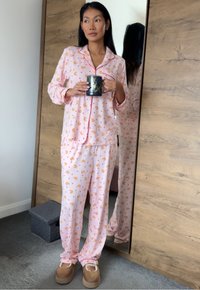Ensemble de pyjama rose avec motif de canetons, comprenant une chemise à boutons et un pantalon assorti. La chemise est bordée d'un passepoil contrastant. Chaussures confortables.