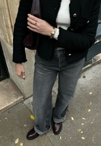 Zwarte textuur blazer met franjes aan de onderkant, witte geribbelde top, grijze wijde jeans en bordeauxrode loafers. Gouden horloge om de pols, clutch tas.
