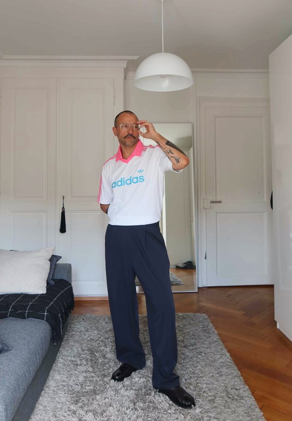 Mężczyzna z wąsami i okularami stoi w pokoju, ubrany w białą i różową koszulkę Adidas, ciemne spodnie i czarne buty, poprawia okulary.