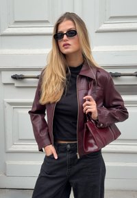 Bordeaux læderjakke med lynlås, båret over en sort bluse. Modellen holder en matchende håndtaske og har sorte brede bukser og solbriller på.