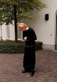 Pulover negru tricotat, pantaloni negri lejeri, curea maro și pantofi negri. Figura stă lângă un copac și o alee de cărămidă texturată.